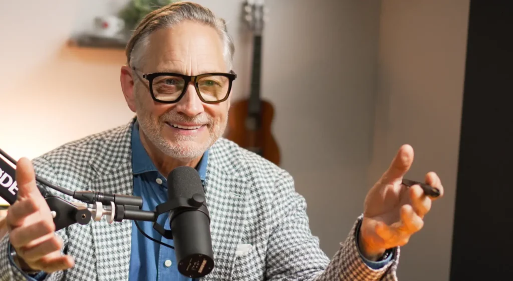 A smiling man with gray hair and glasses, wearing a checkered blazer, gestures while speaking into a microphone during a podcast recording.