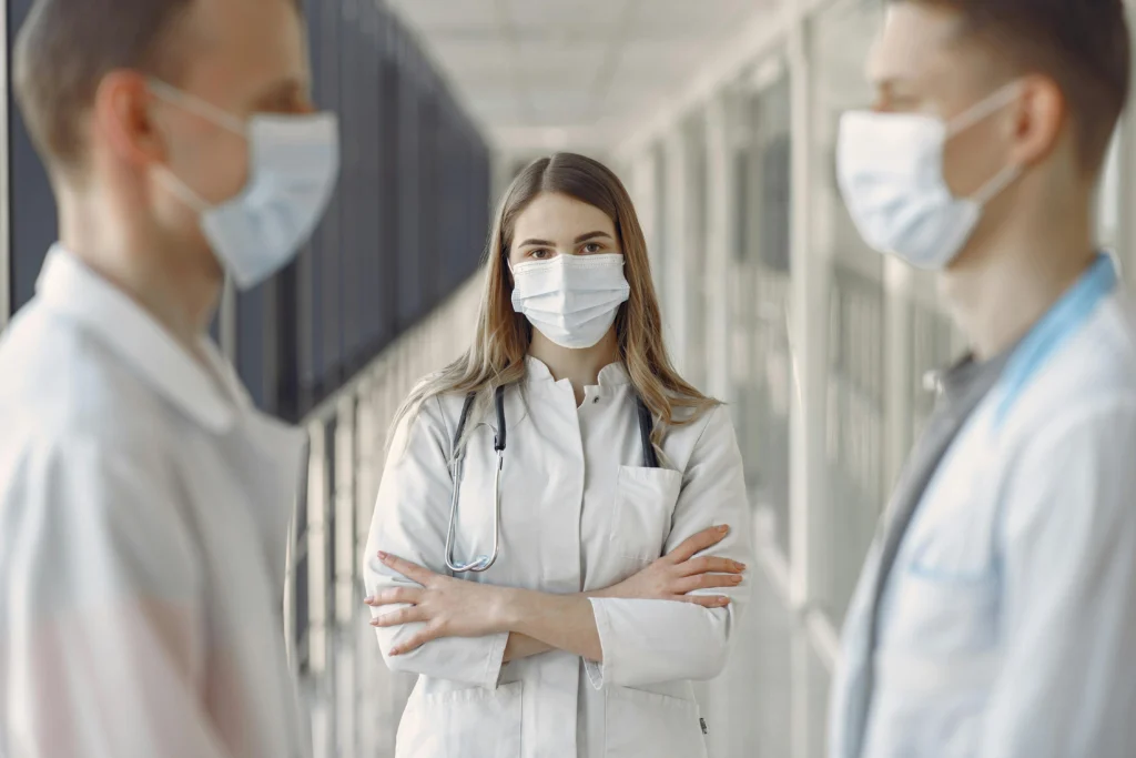 Three medical professionals in white coats and face masks stand in a hallway.
